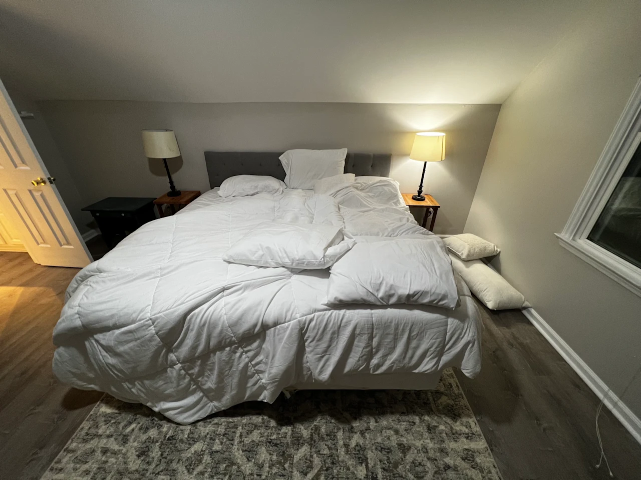An unmade bed with rumpled white bedding and scattered pillows in a softly lit bedroom