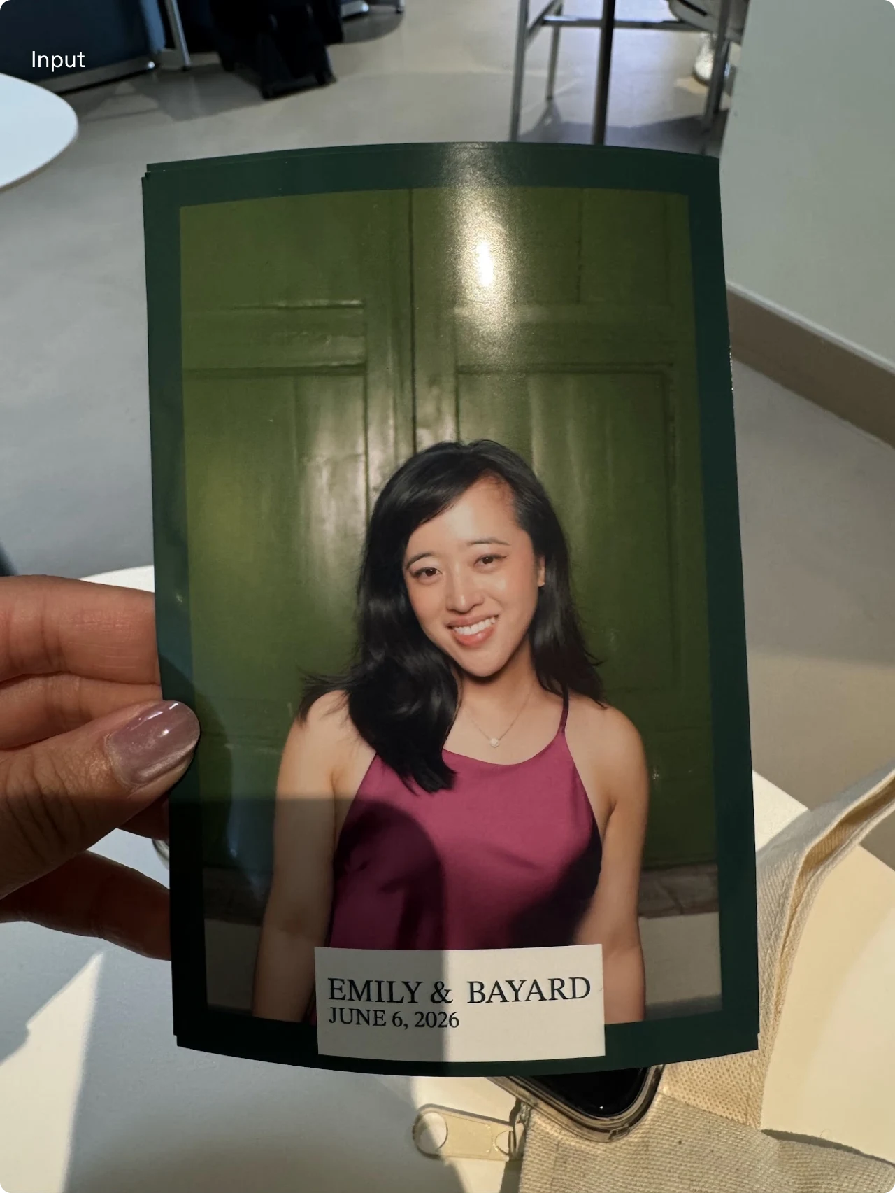 A hand holds a printed photobooth portrait of a smiling woman in a magenta top in front of green doors
